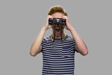 Curious teenage boy looking through binoculars. Shocked teen boy in striped t-shirt using binoculars on gray background.