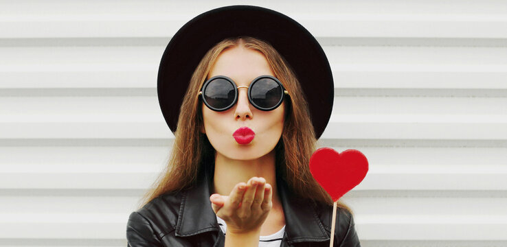 Portrait of beautiful young woman covering her eyes with red heart shaped lollipop blowing red lips sending sweet air kiss on a white background