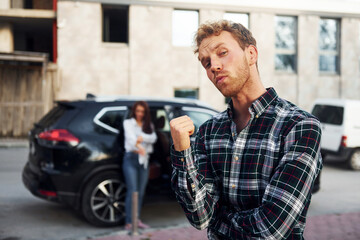 Man in fashionable clothes standing in front of girl that is near the black automobile