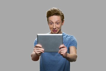 Amazed teen boy looking at digital tablet. Shoked teen guy using pc tablet against gray background.