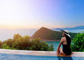 Asian women in black swimsuit and hat siting on side of infinity pool relaxing at sunset time with sea view during vacation retreat. Women happy and enjoy with holiday summer travel time.