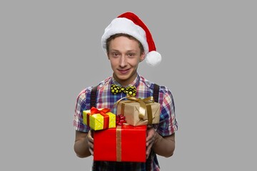 Boy in Christmas hat giving gift boxes. Portrait of handsome teen guy wearing Santa Claus cap handing Christmas present boxes on gray background.