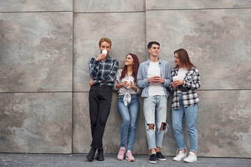 Obraz na płótnie Canvas Group of young positive friends in casual clothes standing together against grey wall with cups of drink in hands