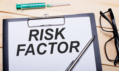 The words RISK FACTOR is written on a white piece of paper next to black-rimmed glasses, a pen and a syringe. Medical concept