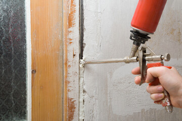 Young adult man hand using spray gun and filling gap with construction foam between old door frame and wall. Closeup. Repair work of home. Renovation process. - obrazy, fototapety, plakaty
