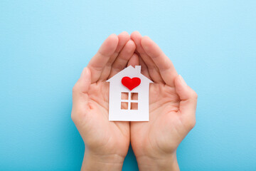 White paper house with bright red heart in young adult woman palms on light blue table background. Pastel color. Closeup. Point of view shot. Top down view. - obrazy, fototapety, plakaty