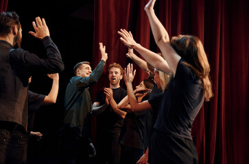 Giving high five, celebrating success. Group of actors in dark colored clothes on rehearsal in the theater - obrazy, fototapety, plakaty