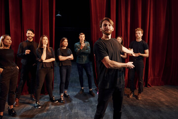 Man standing against people. Group of actors in dark colored clothes on rehearsal in the theater - obrazy, fototapety, plakaty