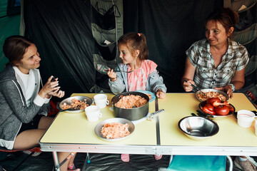 Family having breakfast outdoors on camping during summer vacation. Bread, cottage cheese, cold meat, tomatoes, fruits and coffee cups on table