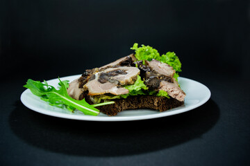 Bitten sandwich with baked meatloaf and herbs, with salad leaves and arugula for a snack on a white plate on black background.