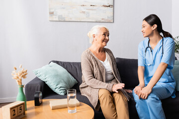 smiling asian nurse and cheerful elderly woman talking on sofa in nursing home, blurred foreground