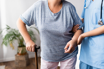 partial view of nurse supporting elderly woman with walking stick in hospital