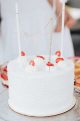 White first birthday cake with white cream and strawberries. Two sticks with triangular flags are inserted. Human silhouette in the background. Summer holiday dessert. Cake with 1 birthday candle