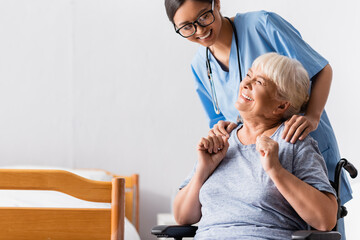 Obraz premium happy asian nurse touching shoulders of aged handicapped woman laughing in wheelchair