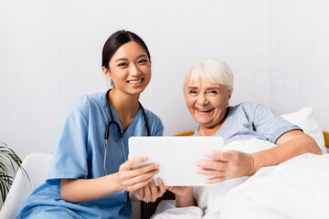 happy asian nurse and laughing senior woman looking at camera while holding digital tablet