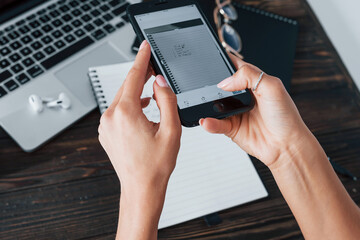 Holding phone. Young female freelancer working indoors in the office at daytime. Laptop and notepad