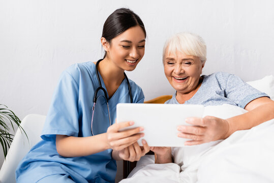 excited asian nurse and cheerful aged woman using digital tablet together