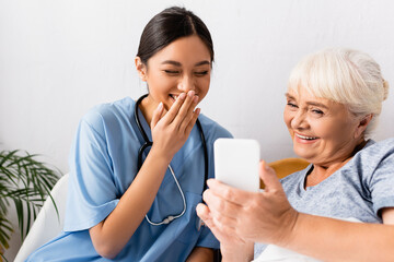 laughing asian nurse covering mouth with hand near cheerful senior woman using mobile phone, blurred foreground