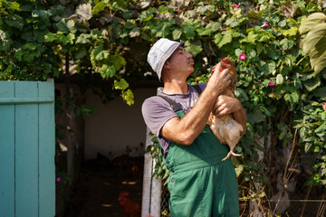 Portrait of happy caucasian farmer holding a brown hen outdoor. Smiling mature man with chicken in...
