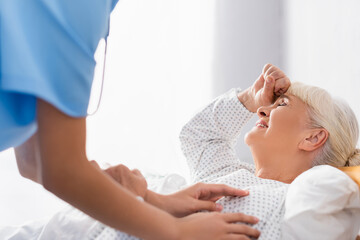Obraz premium nurse touching elderly woman suffering from headache while lying with closed eyes, blurred foreground