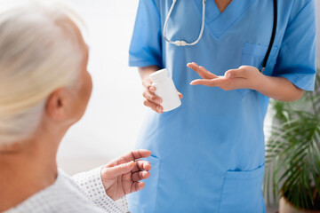 partial view of nurse pointing at container with medicines near aged woman on blurred foreground