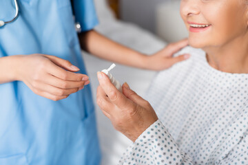 Obraz na płótnie Canvas cropped view of aged, smiling woman holding nasal spray near nurse on blurred background
