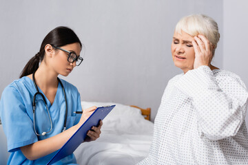 asian nurse in eyeglasses writing on clipboard near aged woman touching head while suffering from migraine