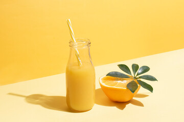A bottle with orange juice and a half of an orange and a straw on a yellow background.