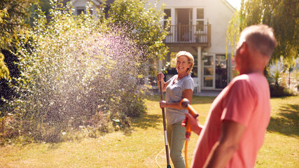 Retired Couple At Work Watering Plants With Hose And Tidying Garden With Rake At Home