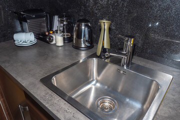Kitchen interior. Kitchen table with sink and grey tile apron on the wall. A table with tea and coffee making facilities. Sink with liquid soap dispenser. Stone countertop on the kitchen table.