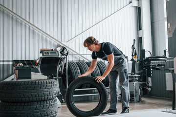 Rolls the tire. Adult man in grey colored uniform works in the automobile salon