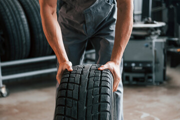 Rolls the tire. Adult man in grey colored uniform works in the automobile salon