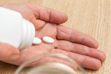 White pills of painkiller or antibiotic for treatment on senior woman hand palm, glass with water, medicines and vitamin supplements concept,close-up view