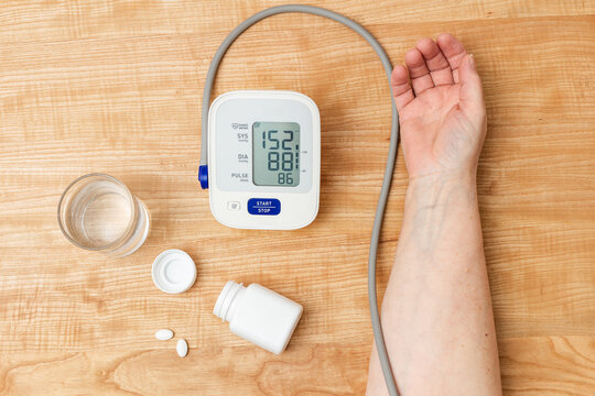 Woman has hypertension, measuring blood pressure on the hand with digital tonometer, hypertensive drugs, treatment pills and water glass on table, top view