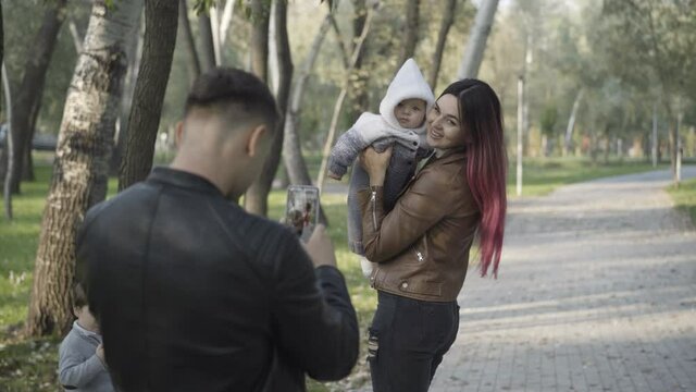 Young Caucasian man taking photos of smiling beautiful woman playing with infant in sunny park. Husband and father enjoying weekend outdoors with wife and little son. Family concept.