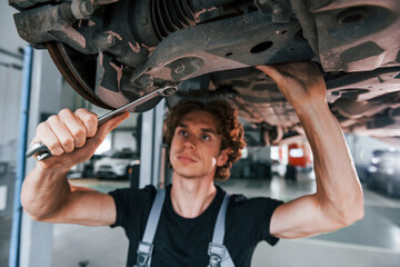 Uses wrench to fix car. Adult man in grey colored uniform works in the automobile salon