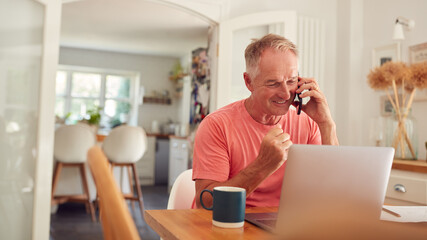 Fototapeta premium Retired Man On Phone At Home In Kitchen Using Laptop Celebrating Good News Or Winning