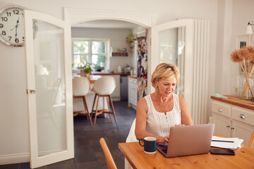 Retired Woman At Home In Kitchen Shopping Online And Managing Domestic Finances Using Laptop