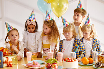 diverse kids in birthday caps celebrate birthday, gathered to congratulate their friend, having...