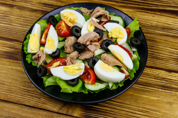 Tasty tuna salad with lettuce, black olives, eggs and fresh vegetables on wooden table