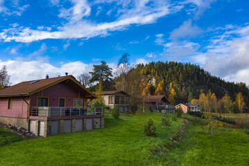 Fototapeta premium Cottage village on the background of a beautiful sky. Rocky mountain with pine trees against a beautiful sky. Wooden houses on a Sunny autumn day. Private houses in the village near the mountain.