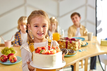 child girl holding birthday cake in hands, kids friends gathered to celebrate her birthday, cute girl enjoys the time with friends, happy