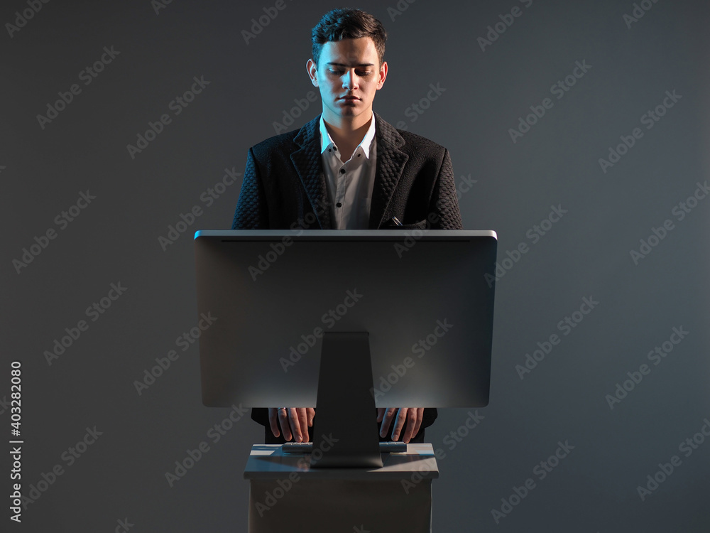 Poster Young man in a business suit is working on computer. Guy is standing and looking the information in computer. Man at a computer on a gray background. Person is working on PC in the middle of frame.