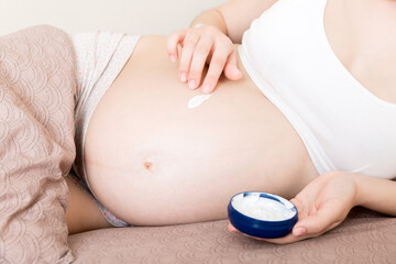a pregnant girl sits at home on the bed and smears smile an anti-stretch mark cream on her stomach. Pregnancy, motherhood, preparation and expectation concept