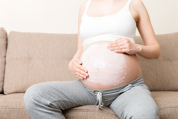 a pregnant girl sits at home on the bed and smears an anti-stretch mark cream on her stomach. Pregnancy, motherhood, preparation and expectation concept