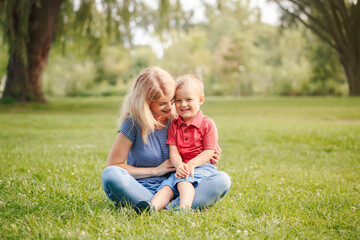 Naklejka na ściany i meble Mothers Day holiday. Young smiling Caucasian mother and laughing boy toddler son sitting on grass in park. Family mom and child hugging having fun together. Happy authentic lifestyle.