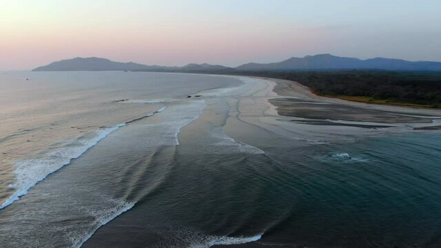 Scenic drone footage of the Pacific Ocean during sunset. Surfers taking surfing lessons in Tamarindo in Costa Rica. Small long waves breaking on the beach. Mountains in the background.