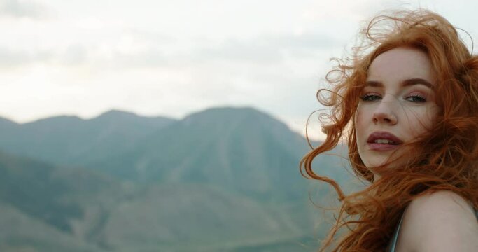 Beautiful female model on top of a mountain posing for photoshoot, reaching her hand out to camera while wind blows her dress and red hair - freedom, tranquility 4k footage