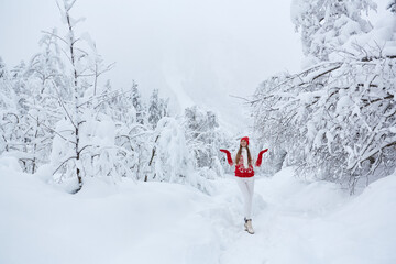 Young caucasian girl in red hat, mittens and knitted sweater on footpath in white snow in a forest of green pines and firs mountain view