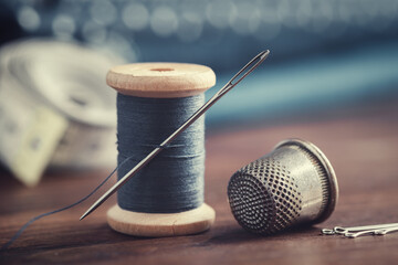 Wooden spool of blue thread, thimble and including pins on table. Blue fabrics for sewing and measuring tape on background. - obrazy, fototapety, plakaty